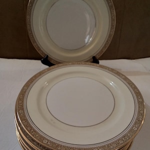 May include: A stack of eight white ceramic dinner plates with a gold rim and a delicate floral pattern. The plates are stacked on a brown surface.