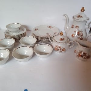 May include: A vintage tea set with a white base and gold trim. The set includes a teapot, sugar bowl, creamer, cups, saucers, and plates. Each piece is decorated with a floral pattern in shades of brown and orange.
