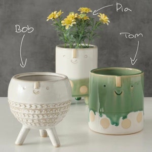 Könnte beinhalten: Drei Keramik-Blumentöpfe mit fröhlichen Gesichtern. Der Topf links ist weiß mit einer strukturierten Oberfläche und drei Beinen. Der Topf in der Mitte ist weiß mit grünen Akzenten und drei Beinen. Der Topf rechts ist grün mit weißen Akzenten. Die Töpfe sind mit "Bob", "Pia" und "Tom" beschriftet.