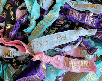 May include: A pile of colorful hair ties with the text "Bride Tribe" printed in gold on them. The hair ties are made of a variety of materials, including elastic, satin, and ribbon.