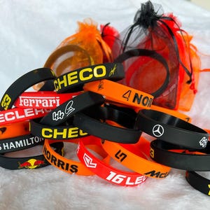 Könnte beinhalten: Eine Sammlung bunter Silikon-Armbänder mit verschiedenen Namen und Logos, darunter "CHECO", "Ferrari", "LECLERC", "4 NOR", "Lewis Hamilton", "Norris" und "16 LE".