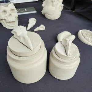 May include: Two white concrete containers with lids, each featuring a small bird sculpture on top. The containers are on a black surface.