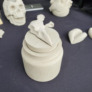May include: A white concrete jar with a lid featuring a bird skull design. The jar is sitting on a black surface.