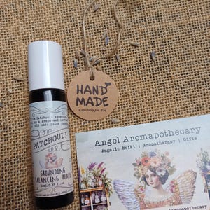May include: A small essential oil bottle with a white cap and a label that reads "Patchouli". A brown tag with "Hand Made" printed on it hangs from a string. A business card with the text "Angel Aromapothecary" is also present.