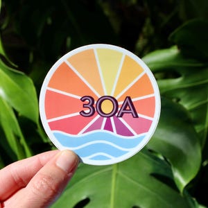May include: A round sticker featuring a colourful sunburst design with the text "30A" in the centre. The sunburst is in shades of orange, yellow, pink and red, with a blue wave at the bottom.