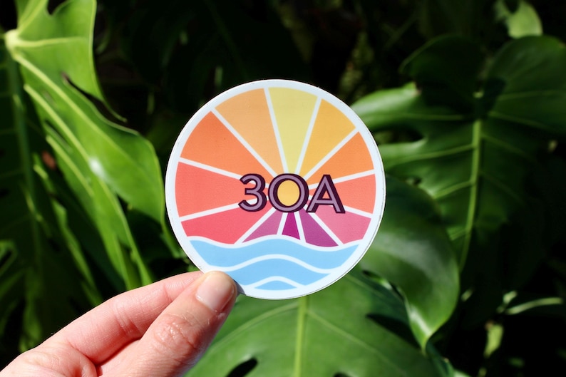 May include: A round sticker featuring a colourful sunburst design with the text "30A" in the centre. The sunburst is in shades of orange, yellow, pink and red, with a blue wave at the bottom.