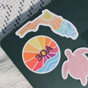 May include: Three colourful stickers on a dark green surface. One sticker is a sunburst design with the text "30A". Another sticker is a pink turtle. The third sticker is a stylised outline of Florida with a sunburst and waves.