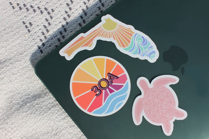 May include: Three colourful stickers on a dark green surface. One sticker is a sunburst design with the text "30A". Another sticker is a pink turtle. The third sticker is a stylised outline of Florida with a sunburst and waves.