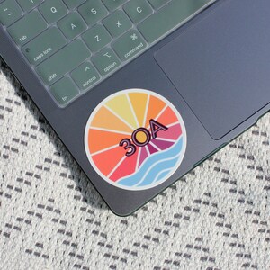 May include: A colourful round sticker with a sunburst design and the text "30A" in a pink outline. The sticker is on a grey laptop.