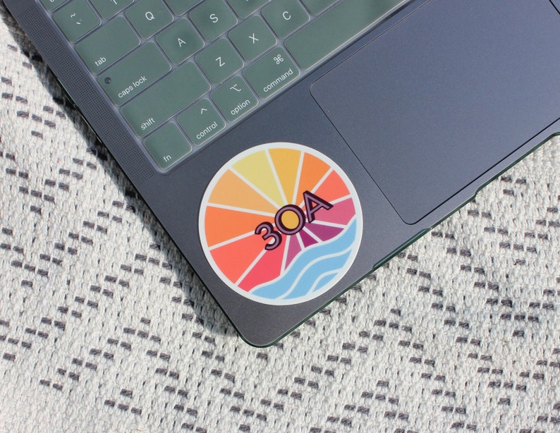May include: A colourful round sticker with a sunburst design and the text "30A" in a pink outline. The sticker is on a grey laptop.