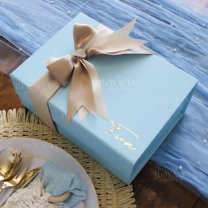 以下が含まれることがあります： 薄い青色のギフトボックスに、金色のリボンと金色の文字で「Tina」と書かれています。ボックスは、薄い青色の生地に白い真珠が置かれています。