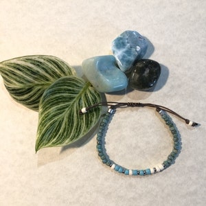 Könnte beinhalten: Ein blaues und weißes Perlenarmband mit einem braunen Kordelverschluss. Das Armband besteht aus kleinen, runden Perlen und ist mit weißen Perlen verziert. Das Armband liegt auf einer weißen Oberfläche mit einem grünen Blatt und blauen und grauen Steinen.