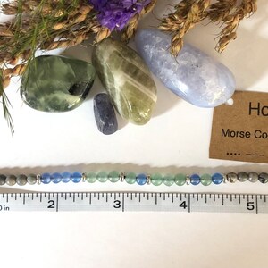 May include: A green, blue, and gray beaded bracelet with silver accents. The bracelet measures approximately 5 inches long. A card with the text "Honor Morse Code Bracelet" is visible in the image.