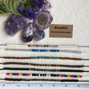 May include: A set of six beaded bracelets with different colors and patterns. The bracelets are laid out on a white surface with a ruler and amethyst crystals. A card with the text "Breathe Morse Code Bracelet" is also visible.