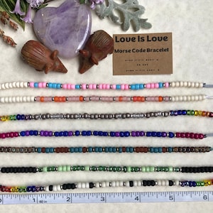 May include: A set of eight colorful beaded bracelets with a ruler for size reference. The bracelets are made with different colored beads and feature a variety of patterns. The bracelets are labeled "Love is Love" and "Morse Code Bracelet".