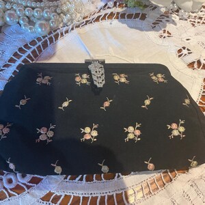May include: Black clutch with embroidered floral details in pink, yellow, and green. The clasp is silver-toned and ornate. The bag is set against a white lace background, with a pearl necklace in the upper left corner.