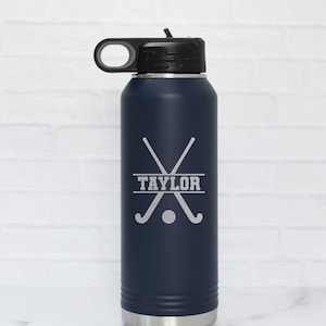 May include: A navy blue stainless steel water bottle with a black lid and a carrying handle. The bottle has a personalized inscription that reads "TAYLOR" with a graphic of two crossed field hockey sticks and a ball.