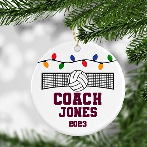 K&ouml;nnte beinhalten: Wei&szlig;er Keramik-Weihnachtsbaumschmuck mit einem Volleyball und Netz, bunten Lichterketten und dem Text "COACH JONES 2023".