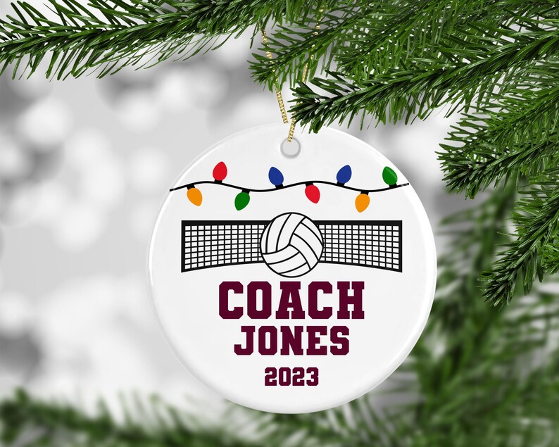 K&ouml;nnte beinhalten: Wei&szlig;er Keramik-Weihnachtsbaumschmuck mit einem Volleyball und Netz, bunten Lichterketten und dem Text "COACH JONES 2023".