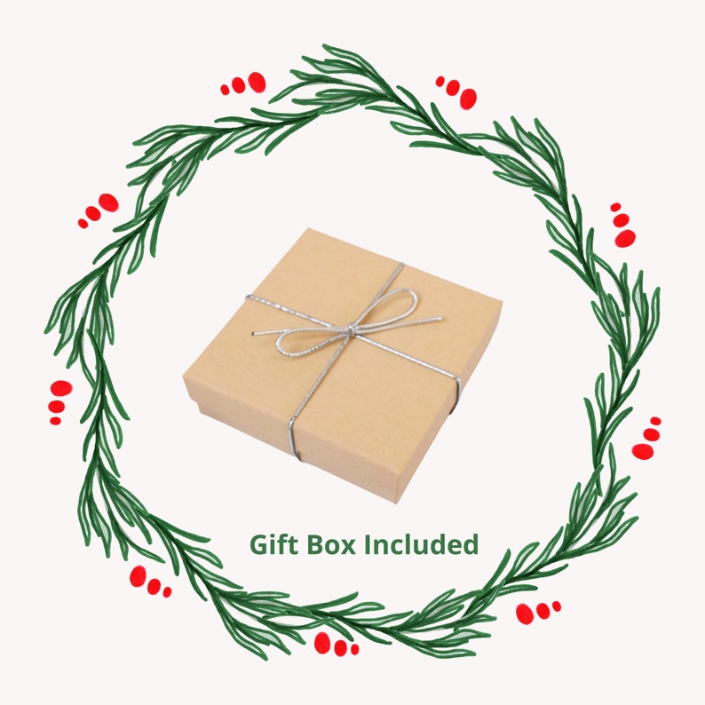K&ouml;nnte beinhalten: Eine braune Geschenkbox mit einem silbernen Band, das zu einer Schleife gebunden ist. Die Box ist von einem gr&uuml;nen Kranz mit roten Beeren umgeben. Der Text "Gift Box Included" befindet sich unter der Box.