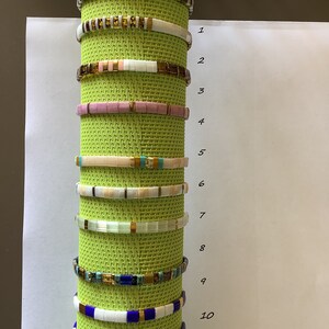 Peut inclure: Dix bracelets colorés faits de petites tuiles carrées. Les bracelets sont empilés sur un tapis vert et numérotés de 1 à 10.