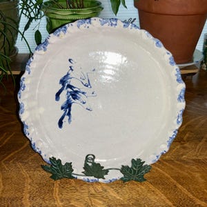 May include: A round ceramic pie dish with a cream-colored base and a scalloped edge featuring a blue floral pattern. The center of the dish displays a hand-painted blue botanical design. The dish is supported by a dark green metal leaf-shaped stand.