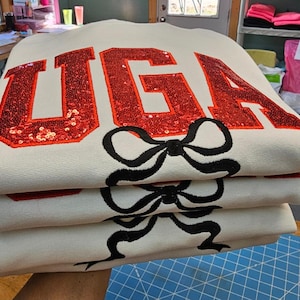 May include: A white sweatshirt with the word "UGA" in red sequins and black bows embroidered on the front. The sweatshirt is folded in a stack.