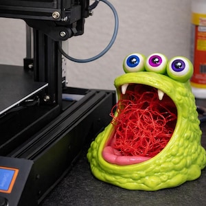 May include: A lime green monster-shaped container with three eyes, white fangs, and a pink tongue. The container is filled with red string. A 3D printer is in the background.