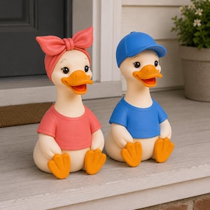 May include: Two duck figurines sitting side by side. The left duck wears a coral t-shirt and a matching coral headband with a bow. The right duck wears a blue t-shirt and a matching blue baseball cap. Both ducks have cream bodies and orange feet.