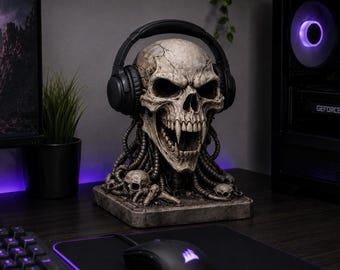 May include: A detailed skull-shaped headphone stand in a gray finish. The skull has a cracked texture, sharp teeth, and is wearing black headphones. The base features smaller skulls and wires. A computer and accessories are in the background.