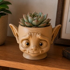 Puede incluir: Una maceta de cerámica beige con forma de criatura de orejas grandes, ojos abiertos y una sonrisa sutil. La maceta presenta una expresión facial detallada con arrugas en la frente y contiene una planta suculenta verde de hojas gruesas.