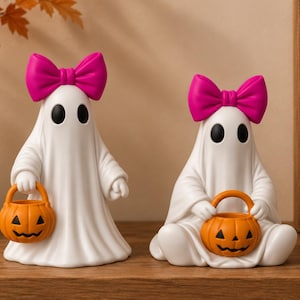 Puede incluir: Dos figuras de fantasmas blancos, cada una con un lazo rosa brillante en la cabeza. Un fantasma está de pie mientras el otro está sentado, y ambos sostienen un pequeño cubo de calabaza de plástico naranja con una cara de jack-o'-lantern negra.