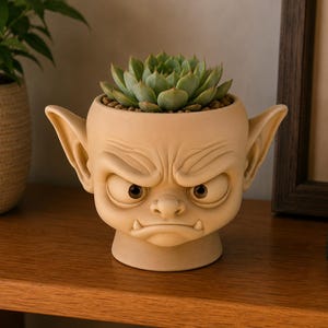 May include: A beige ceramic planter shaped like a grumpy creature with large pointed ears, furrowed brows, and two small fangs. The planter features a round base and contains a green succulent plant with tightly packed, pointed leaves.