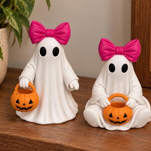Halloween ghost figurine pair with bright pink bows and orange pumpkin trick or treat buckets, featuring one standing ghost and one seated ghost on a wooden shelf.