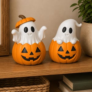 May include: Two decorative figurines featuring white ghosts with black eyes and mouths emerging from orange jack-o'-lantern pumpkins. One ghost wears a small orange pumpkin stem hat, while the other has its arms resting on the pumpkin rim.