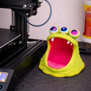 May include: A whimsical, lime green monster-shaped desk organizer with three large, colorful eyes and a wide, open mouth. The interior is bright pink, and the exterior has a textured surface. The organizer sits on a dark surface.