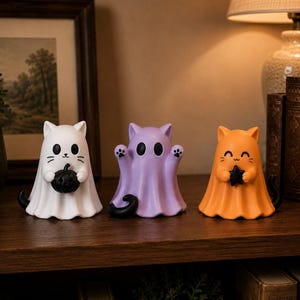 May include: Three cat-shaped Halloween ornaments in white, purple, and orange. The white cat holds a black pumpkin, the purple cat has paw prints, and the orange cat holds a black star. Each has a black tail.
