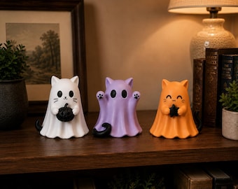May include: Three cat-shaped Halloween figurines in white, purple, and orange. The white cat holds a black pumpkin, the purple cat has paw prints, and the orange cat holds a black star. Each has a black tail.