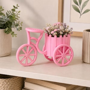 Puede incluir: Una jardinera decorativa en forma de bicicleta rosa con una cesta de estilo valla de madera llena de pequeñas flores rosas y blancas. La bicicleta tiene ruedas grandes y está hecha de un material sólido.