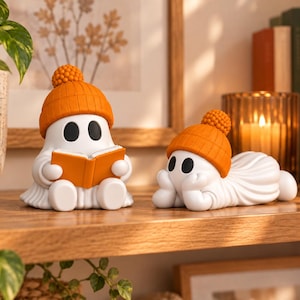 May include: Two white ghost figurines wearing orange knit beanies with pom-poms. One ghost sits upright holding an open orange book, while the other ghost lies on its stomach with its chin resting on its hands. Both have smooth, rounded features.