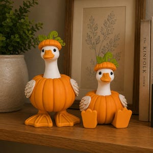 May include: Two white duck figurines with orange pumpkin bodies and green stems on their heads. One duck stands on orange webbed feet, while the other sits with its orange feet extended forward. Both have white wings and orange beaks.