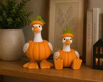 May include: Two white duck figurines with orange pumpkin bodies and green stems on their heads. One duck stands on orange webbed feet, while the other sits with its orange feet extended forward. Both have white wings and orange beaks.