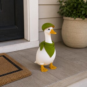 May include: A white duck figurine wearing a green cap and scarf. The duck has yellow feet and a yellow beak. The figurine is standing on a gray surface near a door and a welcome mat.