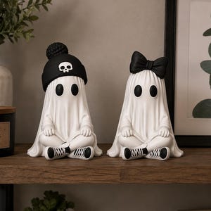Peut inclure: Deux figurines de fantômes blancs assises en tailleur. Le fantôme de gauche porte un bonnet noir avec un emblème de crâne blanc et des baskets noires et blanches. Le fantôme de droite porte un nœud noir sur la tête et des baskets noires et blanches.