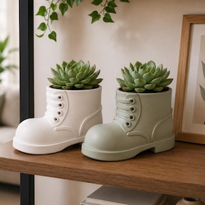 Peut inclure: Deux pots de fleurs en céramique en forme de botte, un blanc cassé et un vert sauge, chacun contenant une plante succulente verte. Les pots ont un design à lacets et sont exposés sur une étagère en bois. Une image encadrée et de la verdure sont en arrière-plan.