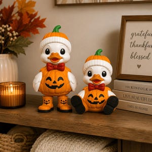 May include: Two white duck figurines dressed in orange pumpkin costumes with green stems on their heads. Each duck wears a white ribbed hat, a red bow tie, and orange pumpkin-shaped boots or feet, featuring black jack-o'-lantern facial expressions.