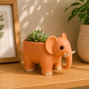 May include: An orange elephant-shaped planter with a succulent plant inside. The planter has large ears, a trunk, and small tusks. The succulent is green and has multiple leaves. The planter is on a wooden surface.