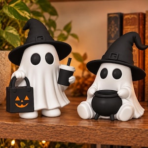Puede incluir: Dos figuras de fantasmas blancos con sombreros de bruja negros. Un fantasma de pie sostiene una bolsa de dulces de calabaza negra y un vaso negro. El otro fantasma sentado sostiene un pequeño caldero negro. Superficies lisas y ojos redondos.