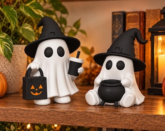 Puede incluir: Dos figuras de fantasmas blancos con sombreros de bruja negros. Un fantasma de pie sostiene una bolsa de dulces de calabaza negra y un vaso negro. El otro fantasma sentado sostiene un pequeño caldero negro. Superficies lisas y ojos redondos.