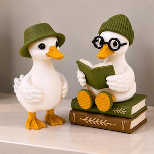 Puede incluir: Dos figuras de patos blancos con picos y patas naranjas. Un pato de pie lleva un sombrero de pescador verde. El otro pato está sentado sobre dos libros apilados, lleva un gorro de punto verde y gafas redondas negras mientras sostiene un libro abierto.